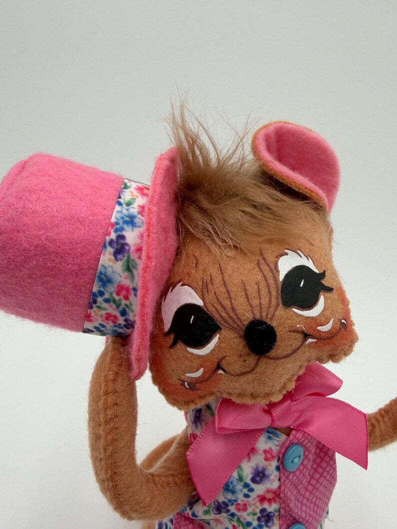 May include: A handmade plush toy mouse with a pink top hat and a floral band. The mouse has brown fur, large black eyes, and a pink bow. The mouse is wearing a pink vest with blue buttons and a floral print dress.