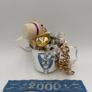May include: A silver bucket with a decorative emblem, filled with a felt figure wearing a hat with a purple band, gold-colored bell, and metallic ribbons. A blue banner with silver text reads "CELEBRATE 2000".