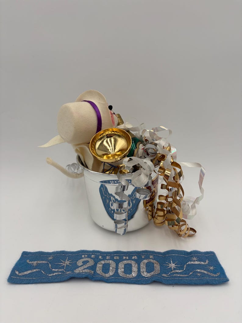 May include: A silver bucket with a decorative emblem, filled with a felt figure wearing a hat with a purple band, gold-colored bell, and metallic ribbons. A blue banner with silver text reads "CELEBRATE 2000".