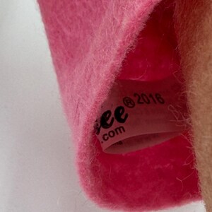 May include: Close-up of a pink plush toy with a tag visible inside. The tag reads "ee .com" and "2016". The plush material is soft and fuzzy. The toy is likely a children's item.