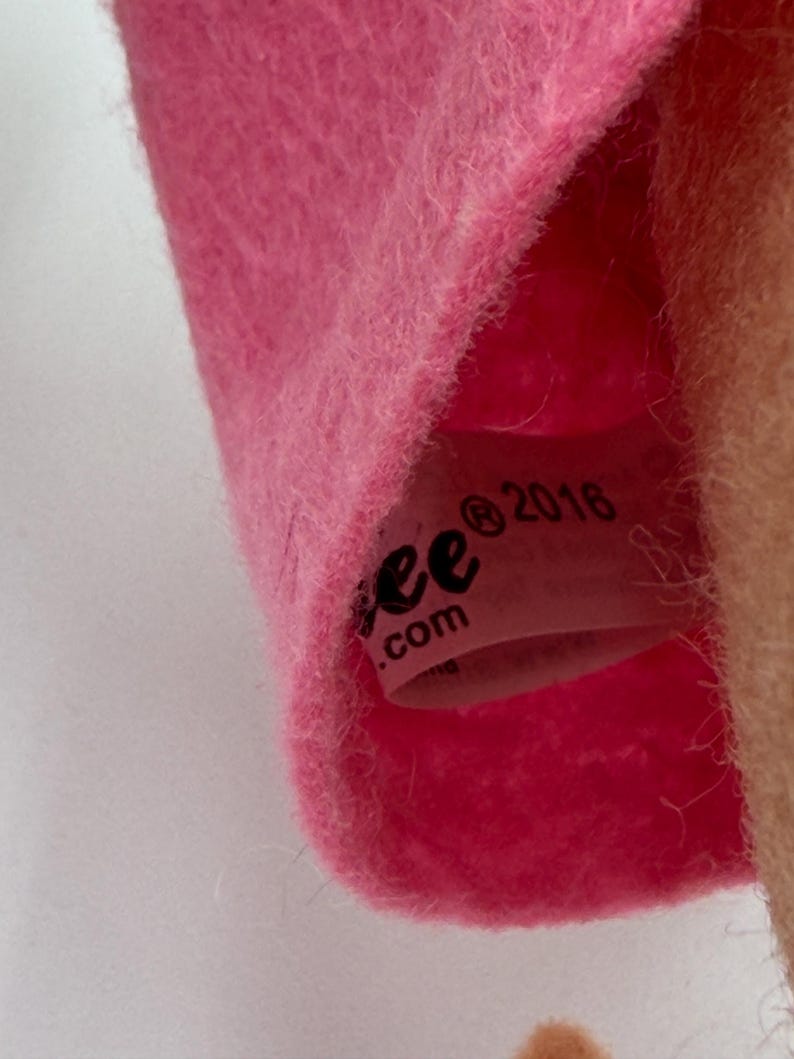 May include: Close-up of a pink plush toy with a tag visible inside. The tag reads "ee .com" and "2016". The plush material is soft and fuzzy. The toy is likely a children's item.