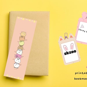 Peut inclure: Un marque-page rose avec une pile de personnages de dessins animés et des étoiles, posé sur un livre marron. Deux étiquettes roses en forme de carte avec le texte "Stray kids" et "skzoo" sont également visibles. Le texte "printable bookmark!" est écrit en dessous.