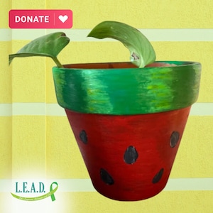May include: A red and green painted terracotta pot with a watermelon design. The pot has black seeds painted on the red portion and a green top with yellow accents. A small green plant is growing in the pot.