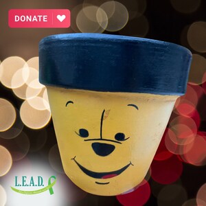 May include: A yellow ceramic flower pot with a black rim and a painted cartoon face of Winnie the Pooh. The pot has a black nose, brown eyes, and a wide smile.