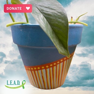 May include: A blue terracotta pot with a yellow and orange striped design and a green plant growing out of it. The pot has the text "L.E.A.D. Let's Empower, Advocate and Do!" on the bottom. The background is a cloudy sky.