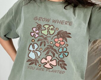Puede incluir: Una camiseta verde con un diseño floral y el texto "Grow Where You Are Planted".