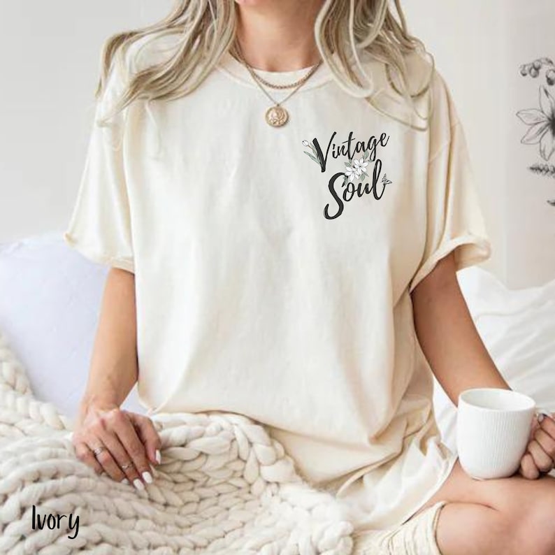 May include: A white t-shirt with the text "Vintage Soul" in black lettering with a small floral design. The shirt is on a white blanket.