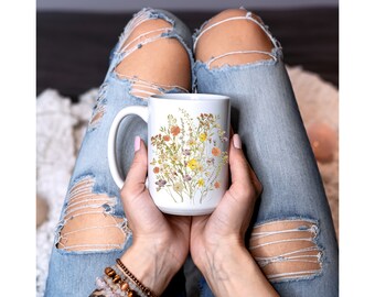 Può includere: Una tazza in ceramica bianca con un design floreale con fiori pressati in tonalità di giallo, arancione e bianco. La tazza è tenuta da una persona che indossa jeans blu.