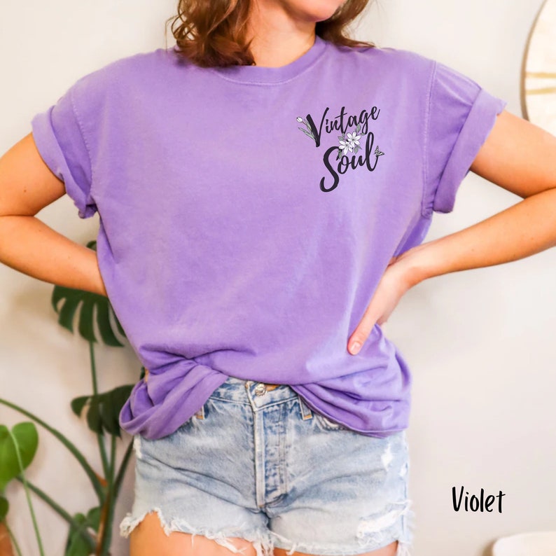 May include: A purple t-shirt with the text "Vintage Soul" and a floral design. The shirt is being worn by a person with blue denim shorts.