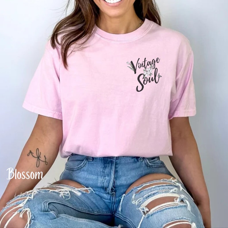 May include: A pink t-shirt with the text "Vintage Soul" and a floral design on the front. The shirt is being worn by a person with a tattoo on their arm.