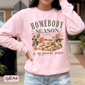 Puede incluir: Una sudadera de cuello redondo rosa claro con el texto verde oscuro "HOMEBODY SEASON" sobre un gráfico de una taza humeante en una manta doblada rodeada de hojas de otoño y bayas, con el texto "is my favorite season" debajo.