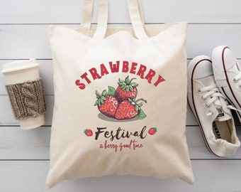 Puede incluir: Una bolsa de lona blanca con texto rojo que dice "Strawberry Festival a berry good time". La bolsa tiene un gráfico de fresas rojas con hojas verdes.