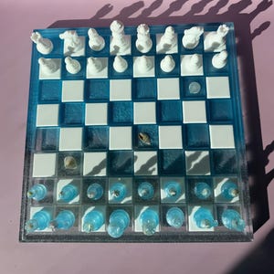May include: A unique chess set with a blue and white checkered board. The board is made of resin and features white chess pieces. The blue squares have a translucent appearance, and some squares contain small seashells. The chess pieces are white.