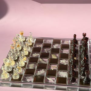May include: A clear acrylic chess set with transparent and black pieces. The chessboard features a mosaic design with botanical elements. The pieces are arranged on a pink background, creating a visually appealing game set.