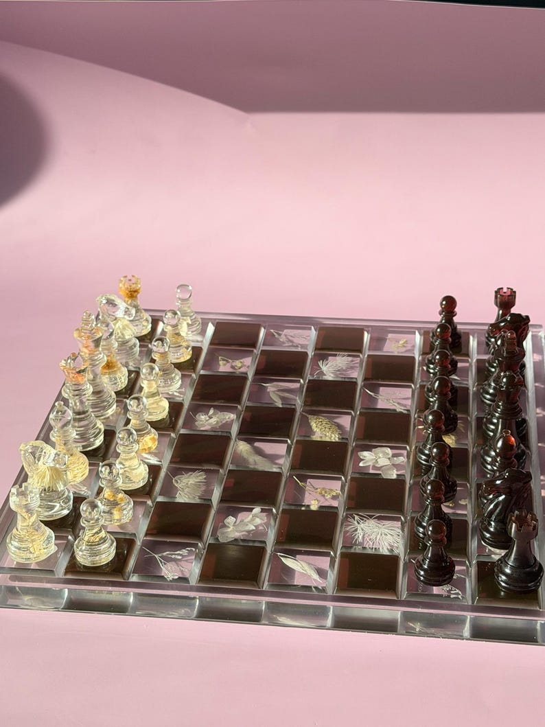 May include: A clear acrylic chess set with transparent and black pieces. The chessboard features a mosaic design with botanical elements. The pieces are arranged on a pink background, creating a visually appealing game set.