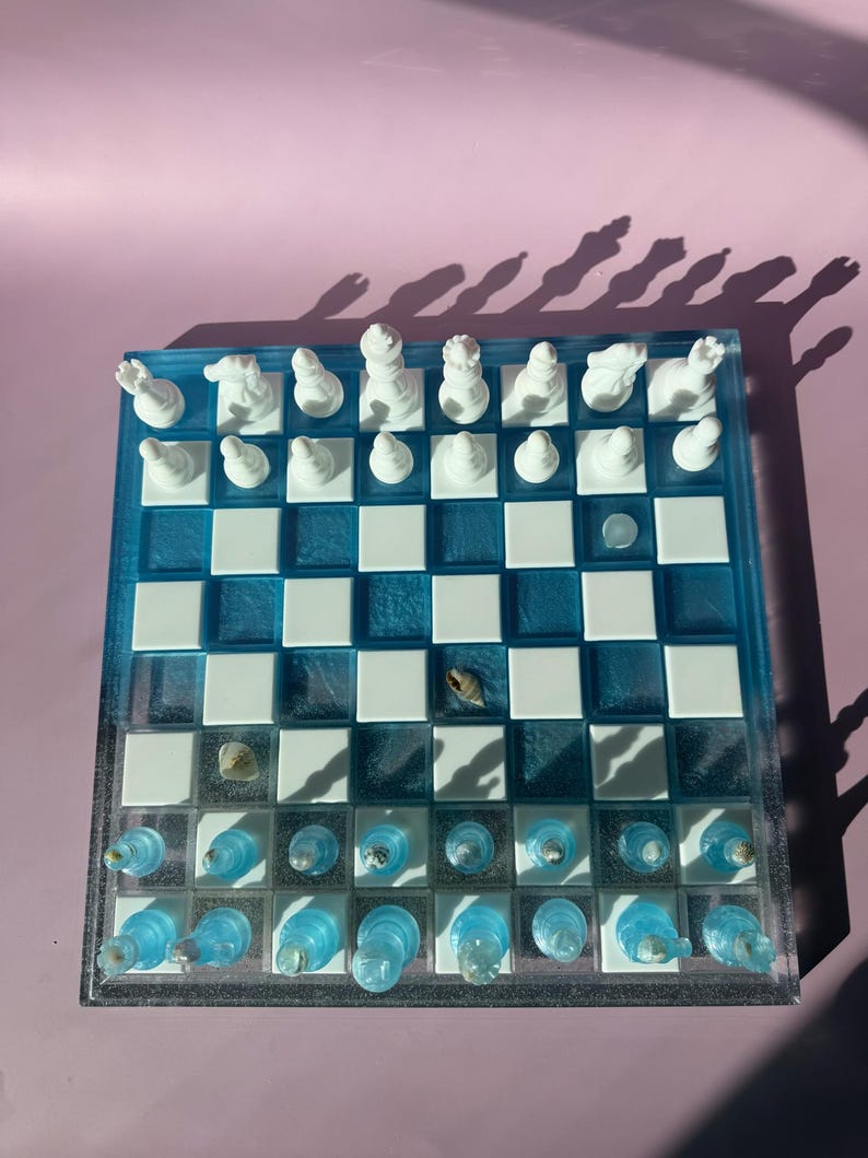 May include: A unique chess set with a blue and white checkered board. The board is made of resin and features white chess pieces. The blue squares have a translucent appearance, and some squares contain small seashells. The chess pieces are white.