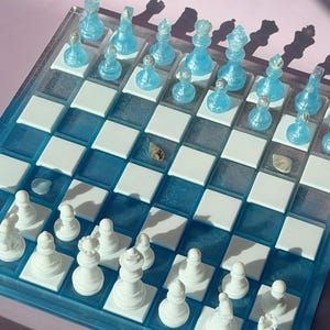 May include: A unique chess set with a clear acrylic board and blue resin accents. The chess pieces are a translucent blue, while the opposing pieces are white. The board features a checkerboard pattern with white squares and blue squares.