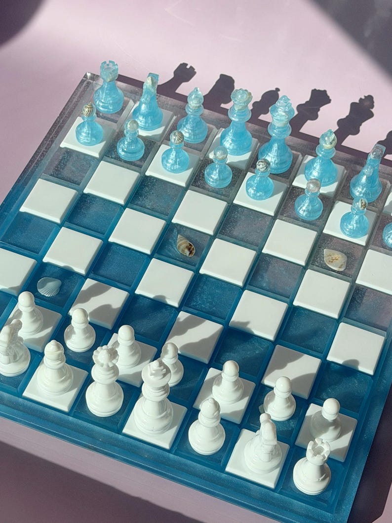 May include: A unique chess set with a clear acrylic board and blue resin accents. The chess pieces are a translucent blue, while the opposing pieces are white. The board features a checkerboard pattern with white squares and blue squares.