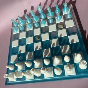 May include: A decorative chess set with a clear, blue-tinted board and white and blue chess pieces. The board has a blue border and white and blue squares. The chess pieces are arranged on the board, ready for play.