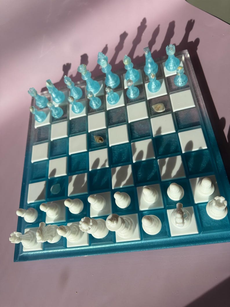 May include: A decorative chess set with a clear, blue-tinted board and white and blue chess pieces. The board has a blue border and white and blue squares. The chess pieces are arranged on the board, ready for play.