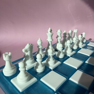 May include: A complete chess set with white pieces arranged on a blue and white checkered board. The pieces include pawns, rooks, knights, bishops, a queen, and a king. The board has a blue resin border and white squares.