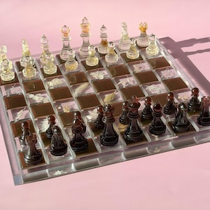 May include: A clear acrylic chess set with transparent and black pieces. The board features a grid of brown and clear squares, some with botanical inclusions. The chess pieces are in their starting positions, ready for a game.