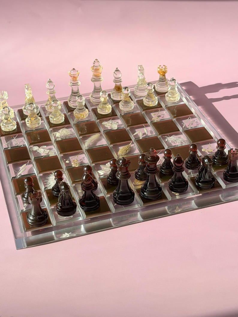 May include: A clear acrylic chess set with transparent and black pieces. The board features a grid of brown and clear squares, some with botanical inclusions. The chess pieces are in their starting positions, ready for a game.
