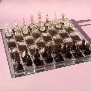 May include: A complete chess set with clear and black pieces on a clear and brown checkered board. The chess pieces are arranged on the board, ready for play. The board is set against a pink background.