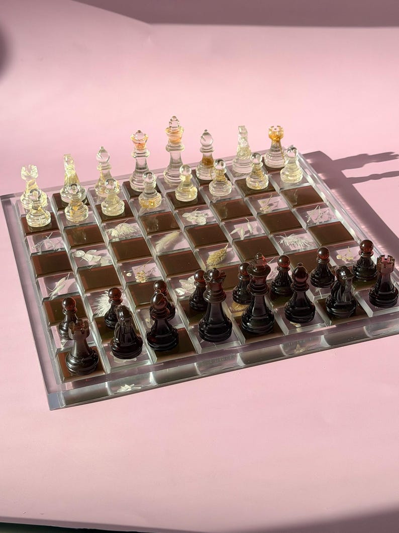 May include: A complete chess set with clear and black pieces on a clear and brown checkered board. The chess pieces are arranged on the board, ready for play. The board is set against a pink background.