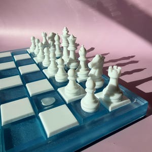 May include: A white chess set with a blue and white board. The chess pieces are arranged on the board, ready for play. The board has a blue resin border and white squares.
