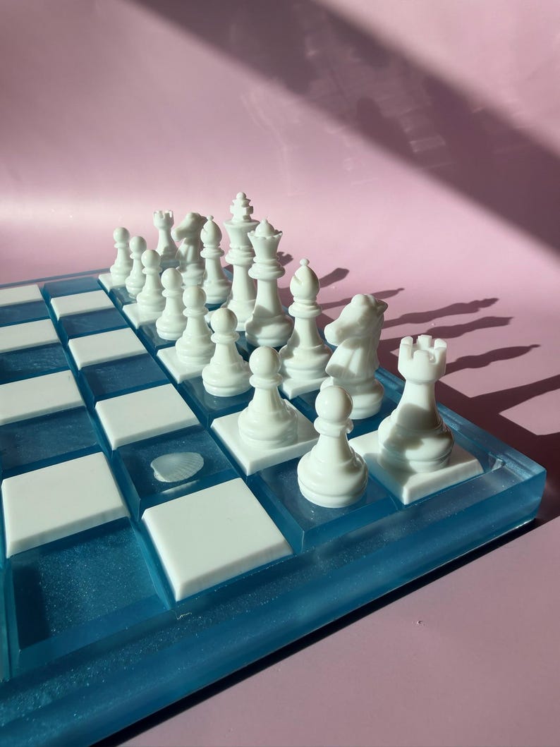 May include: A white chess set with a blue and white board. The chess pieces are arranged on the board, ready for play. The board has a blue resin border and white squares.