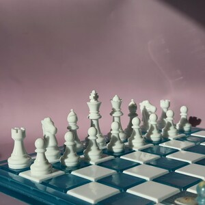 May include: A complete chess set with white pieces arranged on a blue and white checkered board. The pieces include pawns, rooks, knights, bishops, a queen, and a king. The board is set against a pink background.