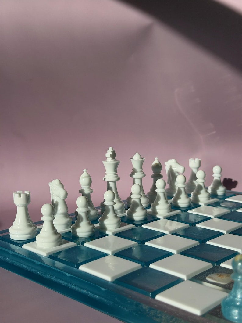 May include: A complete chess set with white pieces arranged on a blue and white checkered board. The pieces include pawns, rooks, knights, bishops, a queen, and a king. The board is set against a pink background.