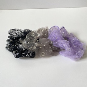 May include: Three scrunchies in black, gray, and lavender. Each scrunchie is made of sheer fabric and adorned with silver star-shaped glitter. The scrunchies are designed for hair accessories.