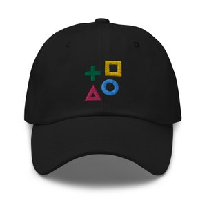 Puede incluir: Gorra de béisbol negra con un diseño bordado de los símbolos del mando de PlayStation: una cruz verde, un cuadrado amarillo, un círculo azul y un triángulo rosa.