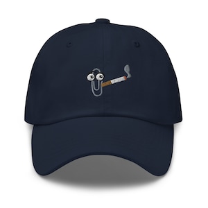 May include: Navy blue baseball cap with an embroidered design of a cartoon paperclip smoking a cigarette. The paperclip has googly eyes and a cigarette in its mouth, with smoke rising from the end. The cap has a curved brim and a fabric strap closure.
