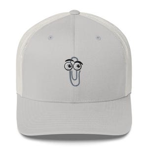May include: A light gray trucker hat with a white mesh back. The front of the hat features an embroidered cartoon paperclip with googly eyes. The hat has a curved brim and a snapback closure.