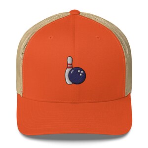 May include: Orange trucker cap with a beige mesh back. The cap has an embroidered bowling pin and ball design in white, red, and navy blue. A fashionable accessory for bowling fans.