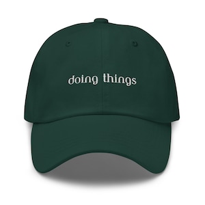 Op de afbeelding: Een donkergroene baseballpet met de tekst "doing things" in wit geborduurd op de voorkant.