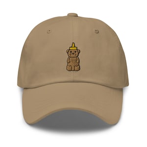 Puede incluir: Una gorra de béisbol de color beige con un oso marrón bordado que lleva una tapa de botella de mostaza amarilla.