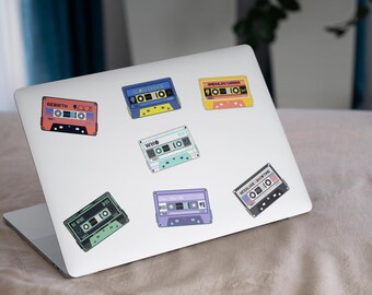 May include: A white laptop with colorful cassette tape stickers. The stickers feature the band names "REBIRTH", "WHO", "METALLICA", "EMERALD GARDEN", "BE MORE", and "INTERLUDE SHORT TAKE".