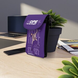 Puede incluir: Funda de tel&eacute;fono morada con una silueta de una persona bailando y el texto "HOPE ON THE STREET" en blanco. La funda tiene una correa negra.