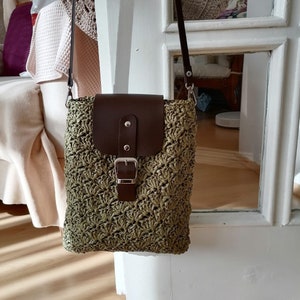 May include: A green crocheted crossbody bag with a brown leather flap and buckle closure. The bag has a long strap and is hanging on a white door.