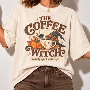 May include: A cream-colored short-sleeve t-shirt featuring a graphic print with the text THE COFFEE WITCH and Fueled by coffee & a little magic. The design includes a coffee mug, a pumpkin, autumn leaves, and a black witch hat with an orange band.