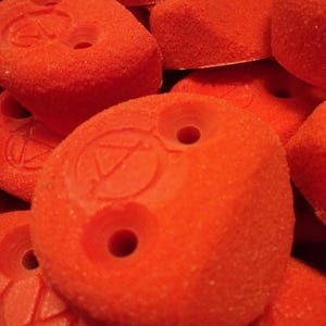 May include: Close-up of a pile of orange climbing holds with a textured surface. The holds have two holes each and a logo with a triangle inside a circle.