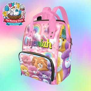 May include: Pink baby backpack featuring a colorful Care Bears design with rainbows, clouds, and the name "Nyemiah". The backpack has a top handle, adjustable straps, and a front zippered pocket. The Nesting Stork logo is in the upper left corner.