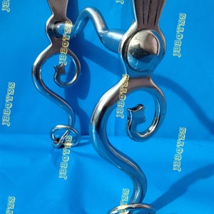 Puede incluir: Un bocado de caballo de acero inoxidable con un diseño curvo. El bocado presenta una articulación central y piezas laterales curvas con remolinos decorativos. La parte superior del bocado tiene una abertura rectangular. El bocado está diseñado para actividades ecuestres.