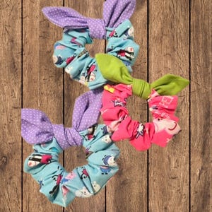May include: Three colorful scrunchies with bow accents. One has a purple polka dot bow, another has a green bow, and the third has a pink bow. The scrunchies have a variety of patterns, including a light blue with cartoon characters.