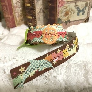 May include: A brown ribbon with a colorful patterned design featuring flowers, zigzags, and a scalloped edge. The ribbon is rolled up and has a green tag attached.