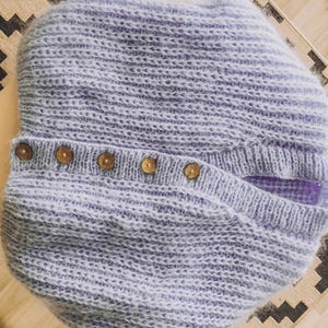 Pu&ograve; includere: Un cappello lavorato a maglia lilla chiaro con patta abbottonata. Il cappello &egrave; realizzato con un filato morbido e strutturato e presenta una fila di cinque bottoni in legno lungo la patta. Il motivo a maglia presenta strisce orizzontali.